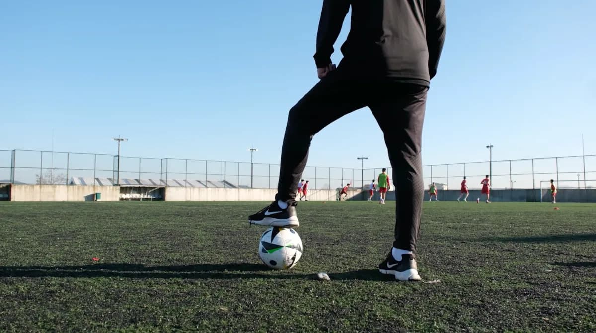 Antrenör ayağını topun üzerine koymuş, arkada saha çalışması sürüyor
